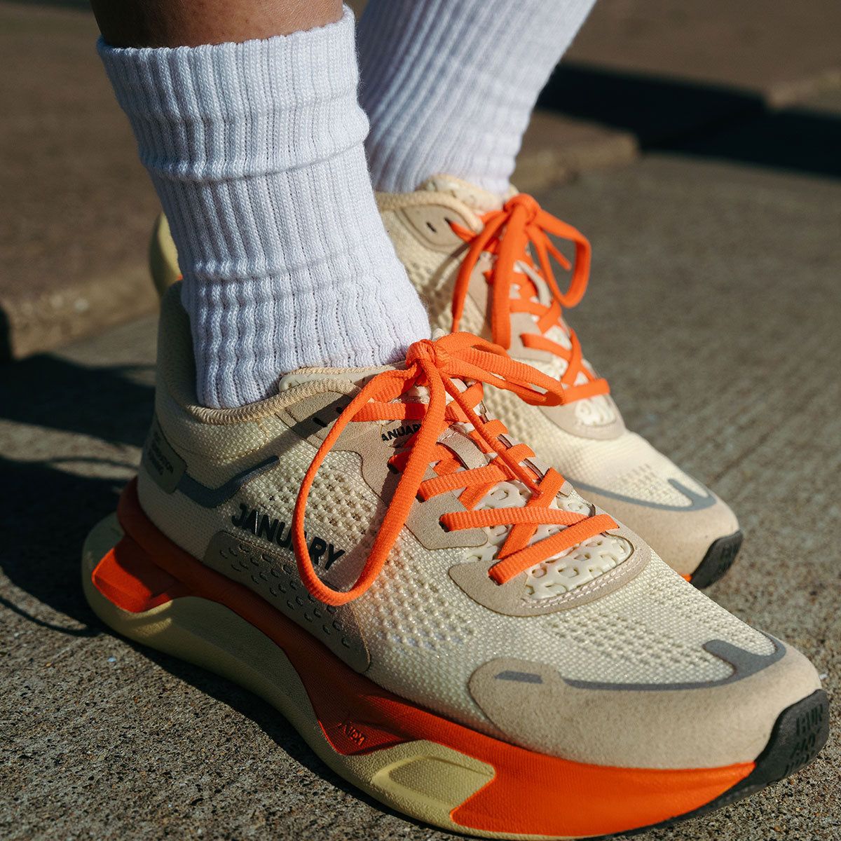 Scignal in Desert Bloom color with orange laces and soles worn with white socks on a concrete surface.
